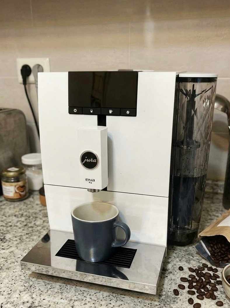 오른쪽에 투명 물통이 달린 흰색 커피머신과 회색 머그잔, 주변에 흩어진 원두