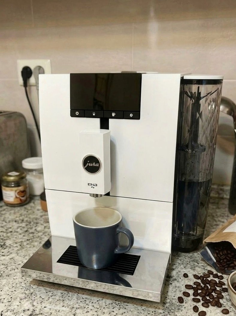 오른쪽에 투명 물통이 달린 흰색 커피머신, 회색 머그잔과 흩어진 원두