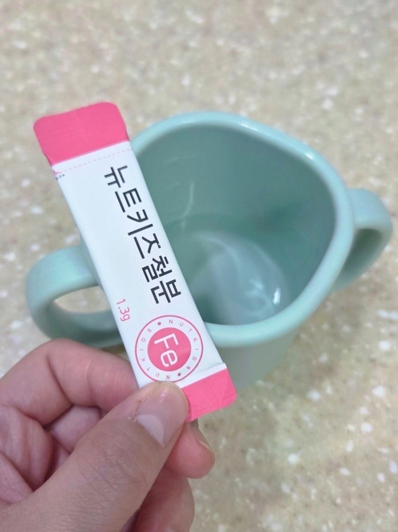 뉴트키즈 철분 스틱 한 포를 든 손과 연두색 아기 컵